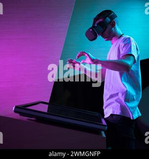 Jeune homme avec un casque VR ayant une expérience de réalité virtuelle immersive dans une pièce avec un éclairage violet, faisant divers mouvements de la main dans les airs. Banque D'Images