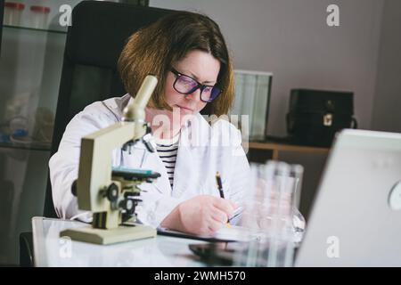 Une femme scientifique caucasienne adulte en lunettes écrit les résultats de son travail. Banque D'Images