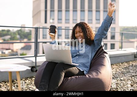 Portrait d'une femme africaine buvant du café tout en levant la main, célébrant les bonnes nouvelles assis sur la chaise de sac de haricots sur la terrasse du toit. Jeune femme Carryin Banque D'Images