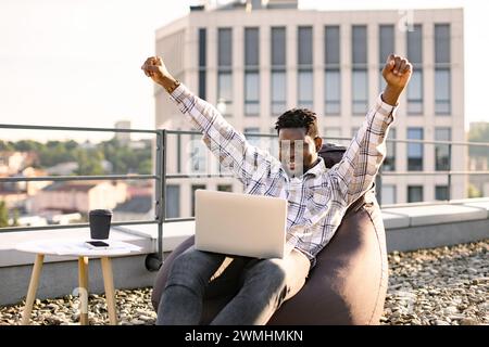 Portrait d'un homme africain buvant du café tout en levant la main, célébrant les bonnes nouvelles assis sur la chaise de sac de haricots sur la terrasse du toit. Jeune homme portant ou Banque D'Images