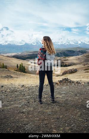 Une voyageuse se tient émerveillée, regardant une vue imprenable sur la montagne, avec l'immensité du paysage qui s'étend devant elle. Son sac à dos et sa tenue vestimentaire suggèrent un voyage dans la nature. Aventurier surplombant Mountain Vista Banque D'Images