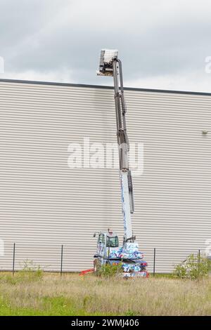 Un ouvrier soulève des matériaux sur le toit d'un entrepôt gris avec un ascenseur hydraulique. Le ciel nuageux laisse entrevoir la possibilité de pluie. Cadre vertical. Banque D'Images