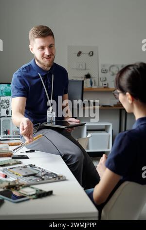 Heureux jeune technicien du service de dépannage utilisant la tablette et regardant une collègue féminine tout en lui expliquant quelque chose Banque D'Images