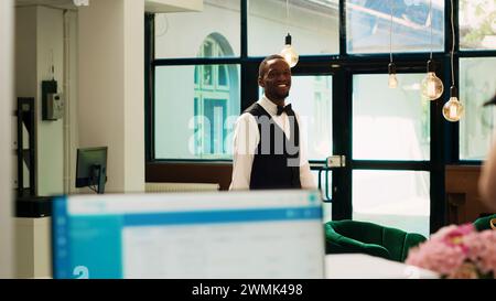 Chasseur de l'hôtel transportant des bagages dans le salon du hall de la réception, travaillant dans le complexe exotique pour aider les clients avec des sacs à roulettes. Concierge professionnel donnant assistance aux clients, service d'enregistrement. Banque D'Images