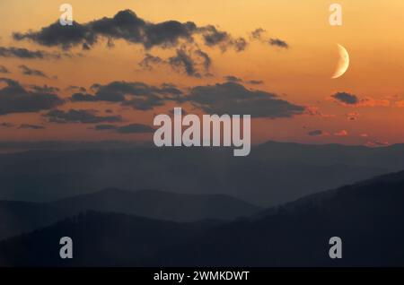 Le croissant de lune brille dans un ciel illuminé au coucher du soleil sur les montagnes Blue Ridge silhouettées ; Fairview, Caroline du Nord, États-Unis d'Amérique Banque D'Images