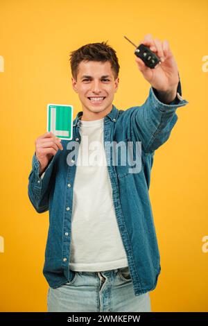 Vertical excité Young Man tenant des clés de voiture et New Driver l signe sur un fond jaune vif. Caucasien Guy célèbre chapeau, il a obtenu un permis de conduire à la couleur de fond. Auto-apprenant. Photo de haute qualité Banque D'Images