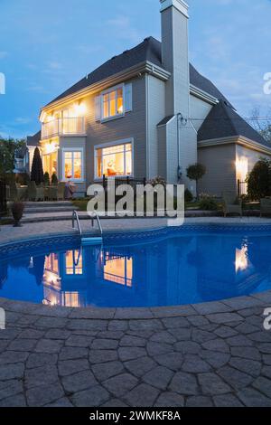 Maison moderne de deux étages avec bardage Havane avec jardin paysagé et piscine creusée illuminée au crépuscule. Banque D'Images