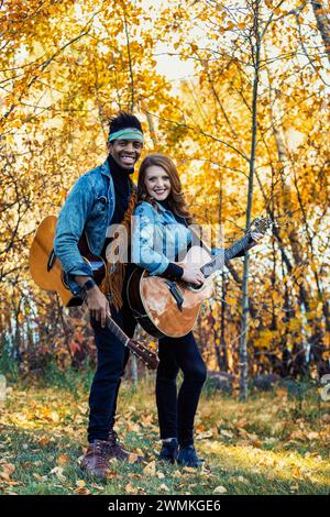 Couple marié de race mixte, souriant et posant pour la caméra tenant des guitares lors d'une sortie en famille d'automne dans un parc de la ville, passer du temps de qualité toge... Banque D'Images