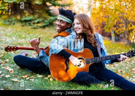 Portrait rapproché d'un couple marié de race mixte assis sur l'herbe dos à dos jouant des guitares avec mari faisant le signe shaka, souriant et ... Banque D'Images