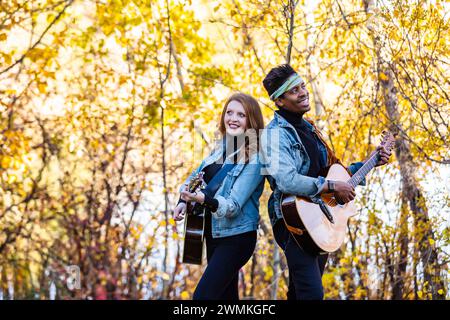 Couple marié de race mixte se tenant dos à dos, jouant de la guitare lors d'une sortie en famille d'automne dans un parc de la ville, passant du temps de qualité ensemble Banque D'Images