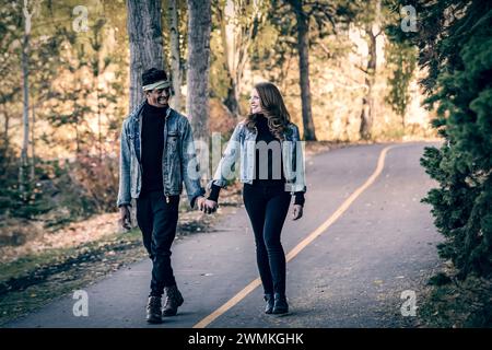 Un couple marié de race mixte se souriant l'un à l'autre et se tenant la main, marchant sur une route dans un parc de la ville lors d'une sortie en famille d'automne, dépensant quali... Banque D'Images