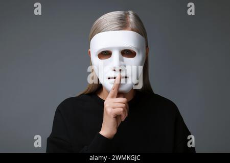 Concept de personnalité multiple. Femme dans le masque montrant le geste de hush sur fond gris Banque D'Images