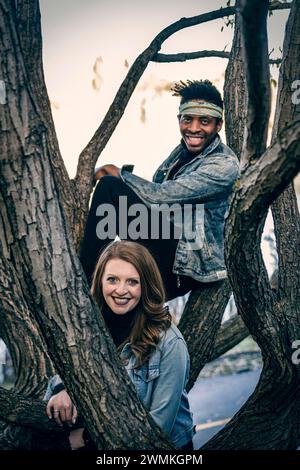 Portrait en gros plan d'un couple de races mixtes souriant et regardant la caméra à travers des branches d'arbres, passant du temps de qualité ensemble lors d'un automne fami... Banque D'Images