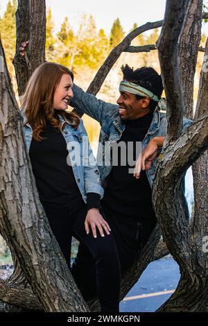 Portrait en gros plan d'un couple de races mixtes souriant l'un à l'autre tout en se reposant sur des branches d'arbres, passant du temps de qualité ensemble lors d'une famille d'automne ... Banque D'Images