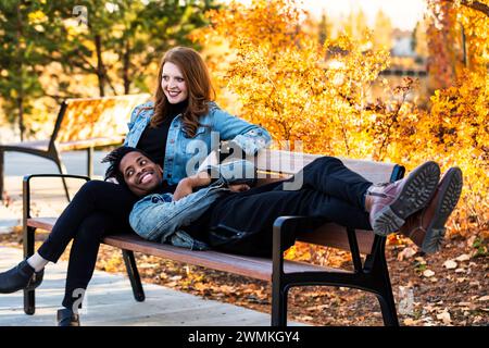 Un couple de courses mixtes souriant et se relaxant sur un banc de parc tout en passant du temps de qualité ensemble lors d'une sortie en famille d'automne dans un parc de la ville Banque D'Images