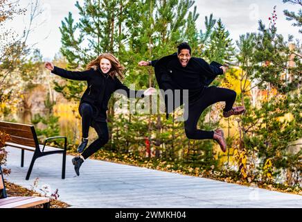 Portrait d'un couple de races mixtes sautant dans les airs, souriant à la caméra et passant du temps de qualité ensemble lors d'une sortie en famille d'automne dans un ci... Banque D'Images