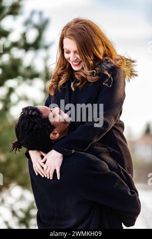 Un couple de races mixtes, mari levant sa femme dans ses bras tout en passant du temps de qualité ensemble lors d'une sortie familiale d'automne dans un parc de la ville Banque D'Images