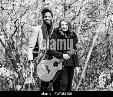 Portrait d'un couple marié de race mixte tenant des guitares acoustiques et souriant à la caméra tout en passant du temps de qualité ensemble lors d'une famil d'automne... Banque D'Images