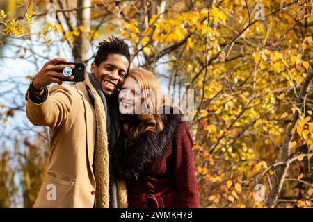 Gros plan d'un couple marié de race mixte prenant un selfie tout en passant du temps de qualité ensemble lors d'une sortie en famille d'automne dans un parc de la ville Banque D'Images