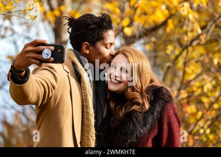 Gros plan d'un couple de races mixtes prenant un selfie ensemble, mari embrassant sa femme sur le front alors qu'elle sourit à la caméra du téléphone portable, tout en dépensant Q... Banque D'Images