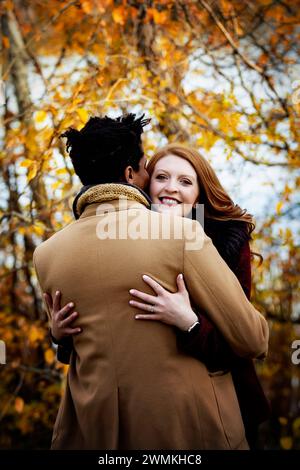 Vue prise de derrière d'un couple de races mixtes s'embrassant avec femme souriant à la caméra, passant du temps de qualité ensemble lors d'un automne fami... Banque D'Images