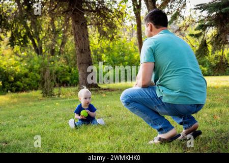 Père passant du temps de qualité et lançant une balle avec son jeune fils atteint du syndrome de Down, dans un parc de la ville pendant un chaud après-midi d'automne Banque D'Images