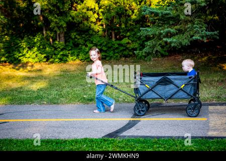 Fille d'âge préscolaire tirant son frère cadet qui a le syndrome de Down dans un wagon sur un chemin dans un parc de la ville pendant une chaude soirée d'automne Banque D'Images