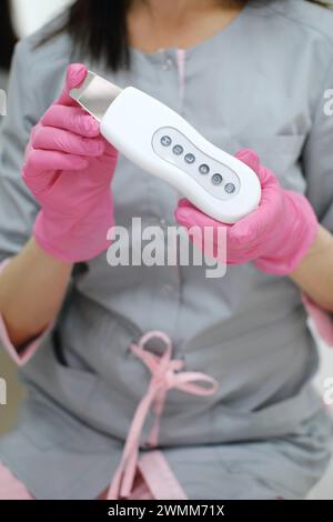 Un cosmétologue en gants roses démontre un appareil de soins de la peau à ultrasons blanc avec des boutons de travail. Photo verticale Banque D'Images