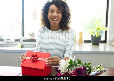 Femme biraciale excitée tenant un cadeau à la maison pendant un appel vidéo de date en ligne Banque D'Images