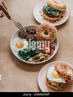 Un bagel aux graines de sésame à moitié mangé, un pâté de saucisse, deux œufs ensoleillés et un sandwich petit déjeuner coupé en deux Banque D'Images