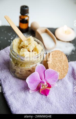 Gommage corporel au sucre fait à la main dans un pot en verre. Bougie brûlante et fleur d'orchidée rose pour la décoration sur fond blanc de studio en pierre noire. Banque D'Images