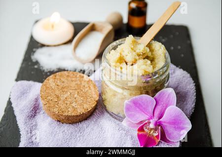 Gommage corporel au sucre fait à la main dans un pot en verre. Bougie brûlante et fleur d'orchidée rose pour la décoration sur fond blanc de studio en pierre noire. Banque D'Images