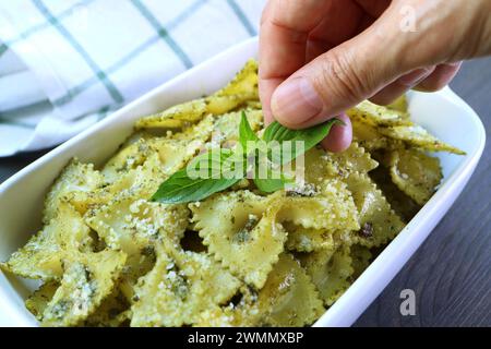 Les gens garnissent le plat de pâtes au Pesto Farfalle fraîchement cuit avec des feuilles de basilic frais Banque D'Images
