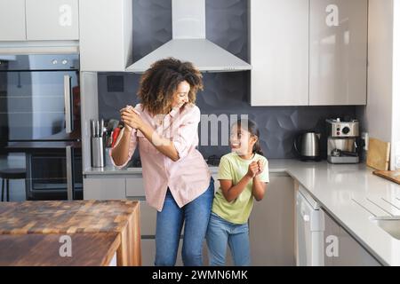 Mère biraciale et fille profiter d'une danse dans une cuisine moderne à la maison. Leur moment spontané ajoute de la chaleur à la maison. Banque D'Images