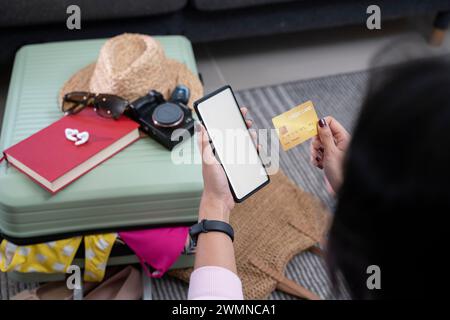 Main de femme prenant une carte de crédit tout en utilisant un téléphone, fond blanc avec une valise dans le fond. Banque D'Images