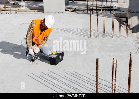 Ouvrier réparateur en uniforme et casque travaillant avec un tournevis sans fil et une boîte à outils. Banque D'Images