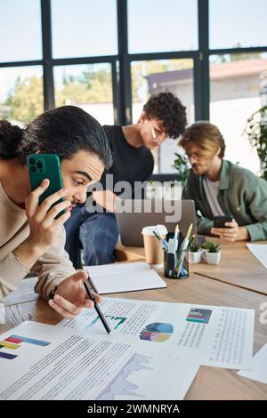 Jeune homme ayant un appel téléphonique et regardant des graphiques à proximité de collègues flous brainstorming sur ordinateur portable Banque D'Images