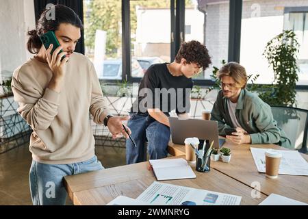 Jeune homme ayant un appel téléphonique et regardant des graphiques à proximité de collègues flous brainstorming sur ordinateur portable Banque D'Images