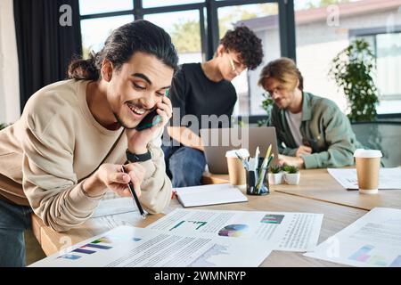 homme heureux ayant un appel téléphonique et regardant des graphiques près de collègues flous brainstorming sur ordinateur portable Banque D'Images