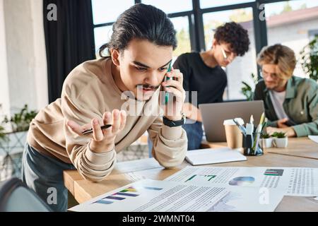 homme concentré ayant un appel téléphonique et regardant des graphiques à proximité de collègues flous brainstorming sur ordinateur portable Banque D'Images