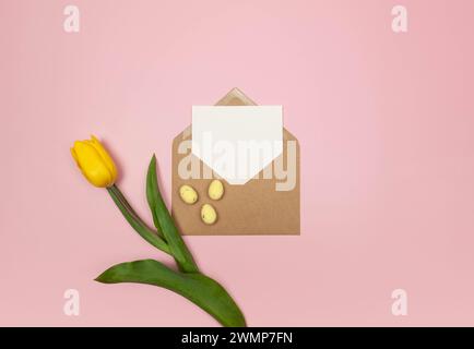 Vue de dessus de tulipe jaune, enveloppe kraft, carte blanche et oeufs de pâques sur fond rose. Composition de pâques de printemps, pose à plat, espace de copie. Banque D'Images