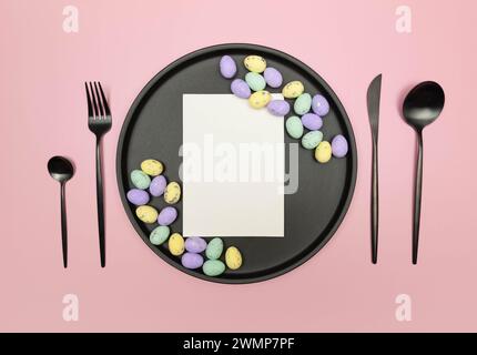 Vue de dessus des oeufs de pâques colorés, carte blanche dans la plaque noire sur fond rose. Table de table, assiette noire et couverts. Composition créative de pâques. Banque D'Images