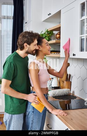 beau couple joyeux et diversifié dans des tenues confortables faisant des tâches ménagères dans la cuisine ensemble et souriant joyeusement Banque D'Images