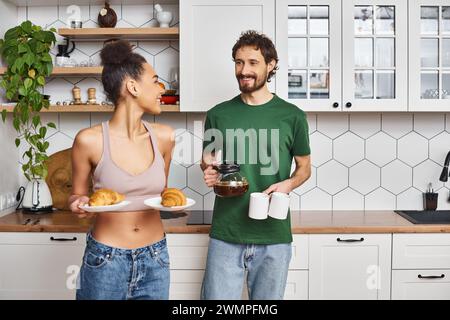 joyeux couple multiracial dans les vêtements de maison se préparant à prendre le petit déjeuner avec du café et des croissants Banque D'Images