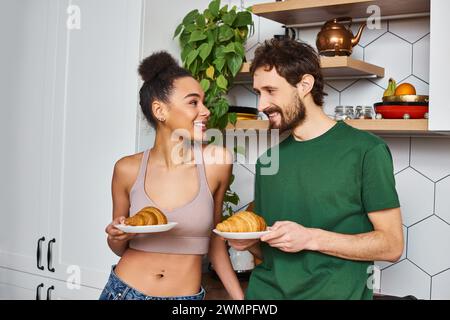 couple jolly interracial dans des tenues décontractées se préparant à manger de délicieux croissants pour le petit déjeuner Banque D'Images