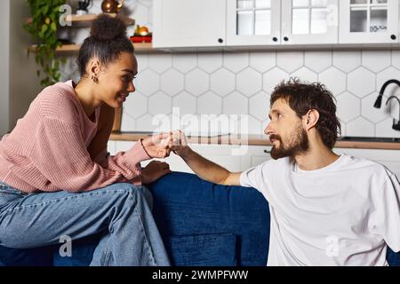 joli couple multiracial joyeux dans des vêtements de maison confortables assis sur le canapé et tenant les mains à la maison Banque D'Images