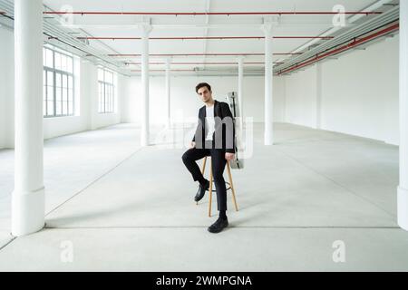 Un jeune garçon est assis sur un tabouret, portant son étui de guitare sur son épaule. Intérieur d'une ancienne usine rénovée. Banque D'Images