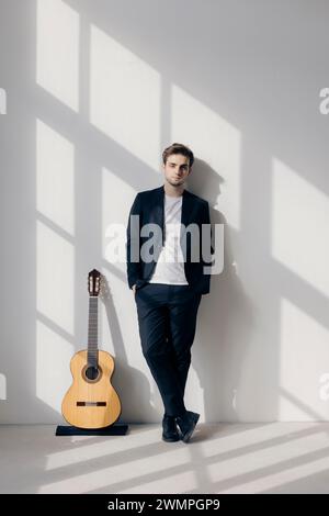 Portrait d'un jeune garçon debout à côté de sa guitare, les mains dans ses poches. Il porte un costume noir. Intérieur d'une ancienne usine rénovée. Banque D'Images