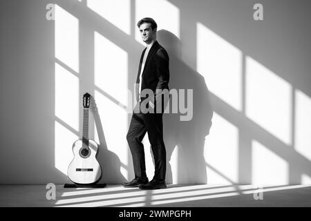 Portrait d'un jeune garçon debout à côté de sa guitare, les mains dans ses poches. Il porte un costume noir. Intérieur d'une ancienne usine rénovée. Banque D'Images