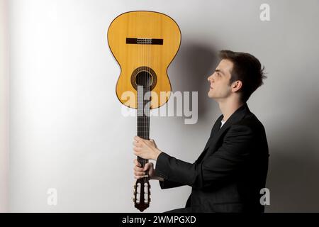 Le jeune garçon tient sa guitare à l'envers et la regarde attentivement. Fond blanc Banque D'Images